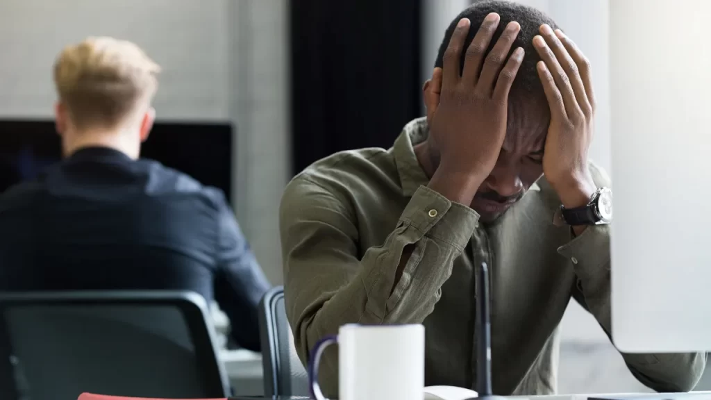 Homem sentado em frente ao computador, segurando a cabeça com as mãos e demonstrando sinais de estresse e cansaço no ambiente de trabalho.