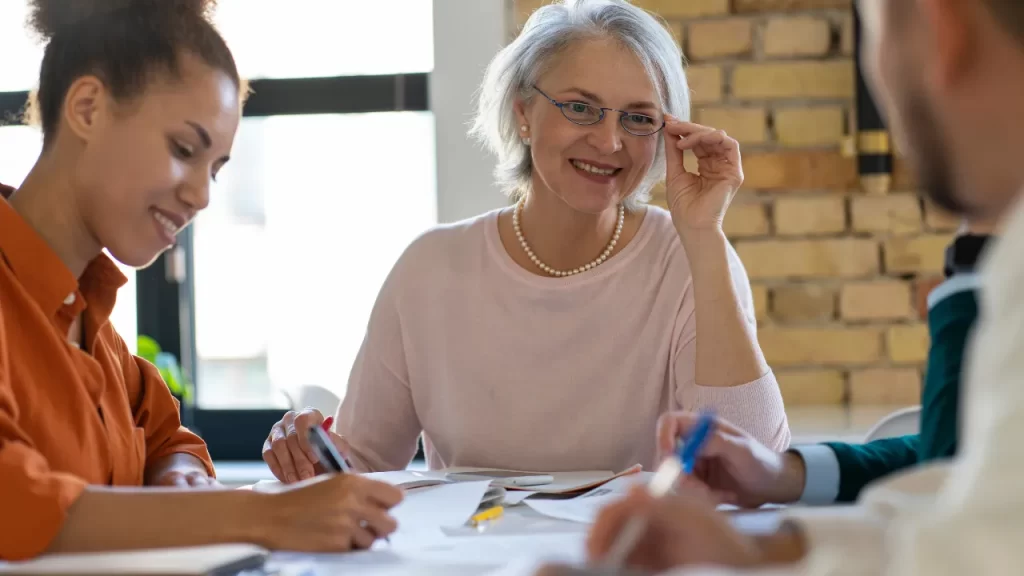 Reunião de equipe com mulher mais velha sorrindo e jovens escrevendo em volta de mesa, simbolizando planejamento de aposentadoria.