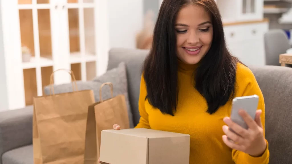 Mulher jovem sorridente segurando uma caixa de encomenda enquanto olha para o celular, representando compras online.