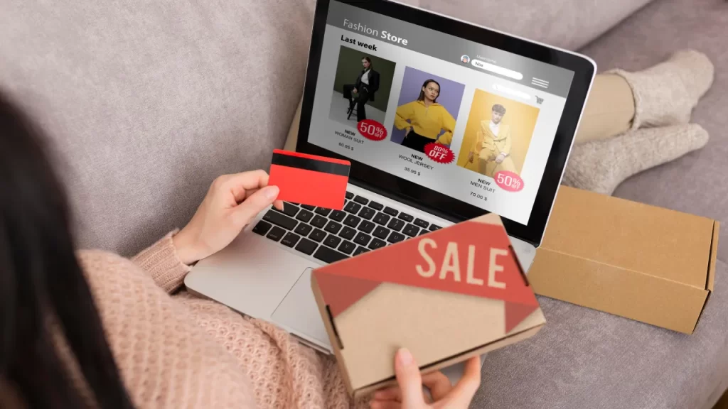 Pessoa segura cartão de crédito e caixa com a palavra "SALE" enquanto realiza compras online em loja de roupas pelo notebook.