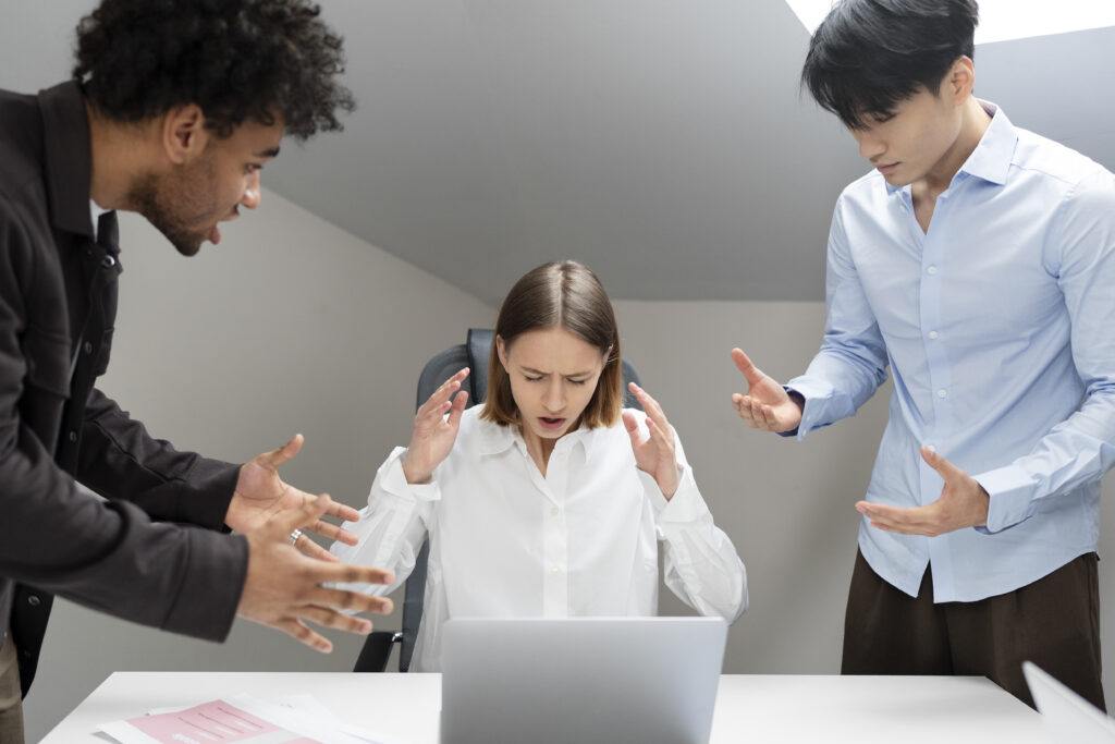 Mulher sentada em frente a um laptop, aparentando estresse, enquanto dois colegas de trabalho em pé gesticulam e falam de forma agressiva ao redor dela.